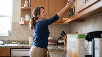 Una mujer con camiseta azul estira el brazo hacia arriba para tomar una caja en un armario de cocina.