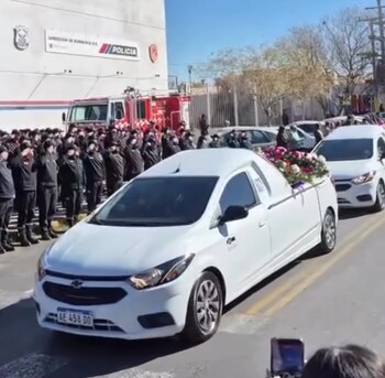 Coche fúnebre blanco con arreglos florales pasa frente a línea de bomberos y policías uniformados que saludan. Edificio de Dirección de Bomberos y Policía