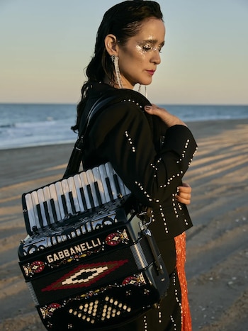 Julieta Venegas, con maquillaje brillante y traje oscuro con perlas, lleva un acordeón Gabbanelli negro en una playa al atardecer, con el mar