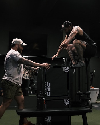 Dos hombres en un gimnasio. Uno de pie sostiene una caja negra. Otro hombre en cuclillas sobre una caja. Cajas con logo "REP FITNESS"