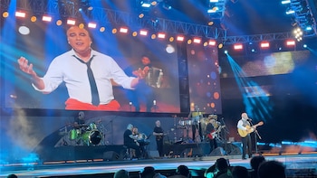 Paz Martínez canta en vivo sobre un escenario, vestido con camisa blanca. Una pantalla gigante detrás proyecta su imagen. Luces azules y naranjas y su banda son visibles