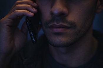 Close de um homem negro latino com barba curta segurando um celular preto próximo à orelha em um ambiente escuro.