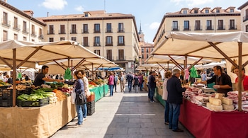 Puestos de venta cubiertos con lonas ofrecen verduras, carnes y quesos a transeúntes en una plaza rodeada de edificios.
