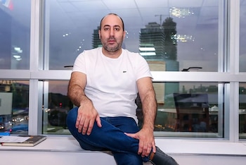 Un hombre de frente con camiseta blanca, pantalón de mezclilla azul y zapatos negros, sentado en elféizar de una ventana con vista a la ciudad