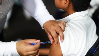 Primer plano de una jeringa azul inyectando el brazo de un niño, cuya mano está ligeramente apretada, mientras una mano adulta sostiene su brazo