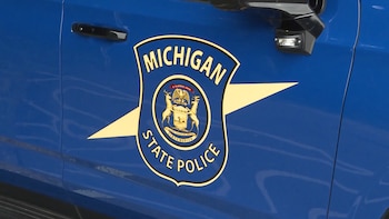 Lateral de vehículo azul con logo de la Policía Estatal de Michigan, escudo dorado y azul, texto Michigan State Police, puerta y espejo retrovisor