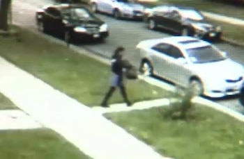 Imagen de baja resolución desde arriba mostrando a una persona caminando en una acera de cemento al lado de una calle. Varios coches, incluyendo uno blanco y uno negro, están en la calle