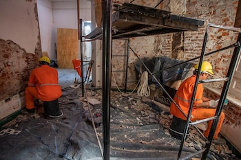 Dos trabajadores con uniformes naranjas y cascos amarillos están sentados en una zona de construcción con paredes de ladrillo expuesto, escombros y andamios