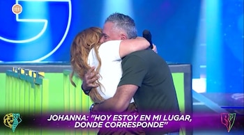 Una mujer con cabello largo rojizo-castaño abraza a un hombre de cabello canoso en un estudio de televisión con luces azules y verdes