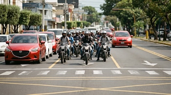 Un grupo de motocicletas con conductores avanza en una calle con líneas viales pintadas, entre filas de automóviles detenidos. Hay edificios y árboles al fondo.