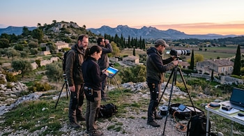 Cuatro personas con equipos fotográficos, prismáticos y una tableta en una colina frente a un valle con casas de piedra y montañas al amanecer.