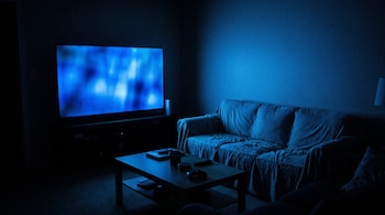 Una habitación oscura con un televisor encendido de pantalla azul, un sofá cubierto con una manta y una mesa de centro con libros, un vaso y un control remoto.