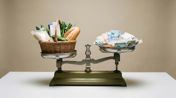 Balanza antigua con canasta de arroz, leche, pan y verduras en un lado y billetes de pesos dominicanos en el otro, inclinada hacia los productos.