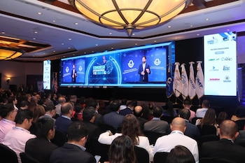 Auditorio con asistentes, escenario con gran pantalla LED mostrando un orador, banderas en el escenario, pantalla lateral con logotipos de patrocinadores, luces de techo