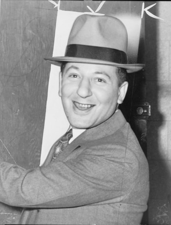 Retrato en blanco y negro de un hombre sonriente con un sombrero de fieltro y traje. Mira directamente a la cámara mostrando los dientes. Fondo oscuro y liso