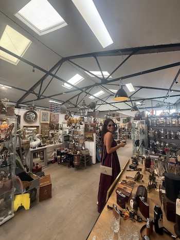 Pampita, con vestido largo y gafas de sol, sonríe en una tienda de antigüedades con techos altos, tragaluces y estantes llenos de objetos