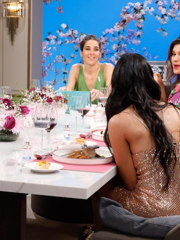 Daniela Celis, vestida de verde, sonríe sentada a una mesa decorada con flores y copas de vino; otras dos mujeres parcialmente visibles la acompañan