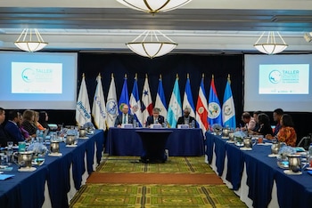Sala de conferencias con una larga mesa azul, sillas, pantallas, tres hombres en la mesa principal y banderas de países centroamericanos
