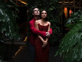 Hombre y mujer abrazados, ambos de pie y mirando hacia arriba, rodeados de abundante vegetación verde, plantas grandes, hiedra y faroles iluminados