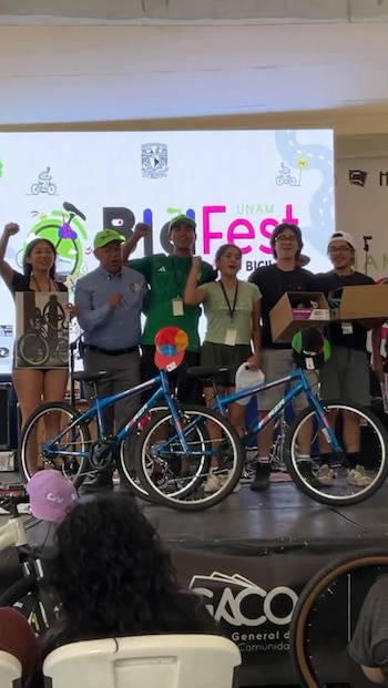 Seis personas celebran en un escenario frente a un cartel de "UNAM Bici Fest", con dos bicicletas de montaña azules y cascos de colores en primer plano
