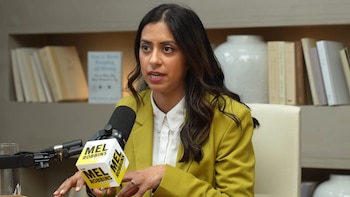 Una mujer con blazer verde lima y camisa blanca sentada en un escritorio de madera, hablando por un micrófono con el logo de The Mel Robbins Podcast