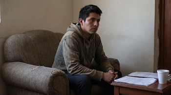 Un joven con cabello oscuro, vestido con una sudadera con capucha, se sienta en un sofá gastado, con las manos entrelazadas, mirando hacia la derecha.