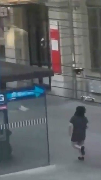 Vista borrosa de una persona corriendo de espaldas en un área urbana o estación de tren, con un cartel azul y edificios al fondo