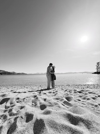 Imagen en blanco y negro de una pareja abrazándose en una playa arenosa junto a un lago, con montañas nevadas al fondo y un sol brillante