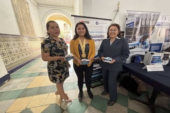 Tres mujeres de pie sostienen cajas pequeñas frente a paneles informativos y una mesa con instrumental médico en un pasillo con azulejos