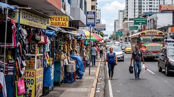 Puestos de venta en una acera, peatones en la calzada, vehículos en marcha y un autobús multicolor en una calle de la ciudad de Panamá.