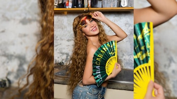 Shakira, con cabello largo y rizado, sonríe a la cámara mientras se ajusta unas gafas de sol y sostiene un abanico verde y amarillo con la palabra 'Loba'