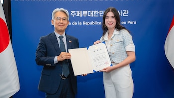 Dos personas, un hombre y una mujer, sostienen un diploma. Detrás, una pared azul con el nombre de la Embajada de Corea del Sur en español y coreano