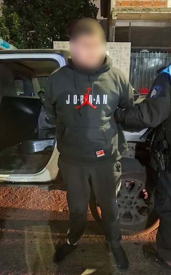 Un hombre con rostro borroso es sujetado por un agente de policía junto a un vehículo policial blanco con puerta abierta, en una calle de noche