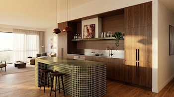 Una moderna cocina con una isla de azulejos verdes y taburetes, gabinetes de madera oscura, se conecta a una sala de estar con sofá y ventana grande.