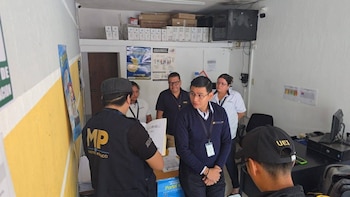 Un agente del Ministerio Público, de espaldas, revisa un documento frente a un funcionario de DENAP en una oficina con estantes y computadoras, mientras otros tres observan la escena