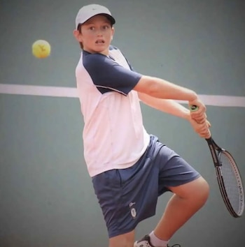 Un niño con gorra blanca, camiseta blanca y azul, y pantalones cortos azules, golpea una pelota de tenis amarilla con una raqueta en una cancha