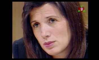 Primer plano del rostro de una mujer con cabello castaño oscuro y piel clara. Tiene una expresión seria con arrugas en la frente. Un logo "9" en su pelo