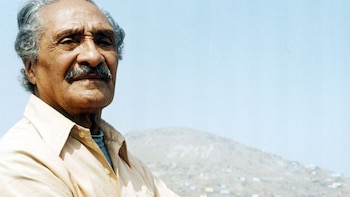 Primer plano de un hombre anciano de tez oscura, cabello canoso y bigote, con camisa clara. Detrás, un cerro árido con casas y la palabra 'COMAS' escrita