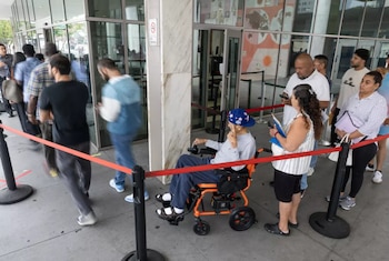 Fila de personas adultas en el exterior de un edificio. Un hombre con gorra en silla de ruedas eléctrica se ubica detrás de una cinta roja junto a otras personas