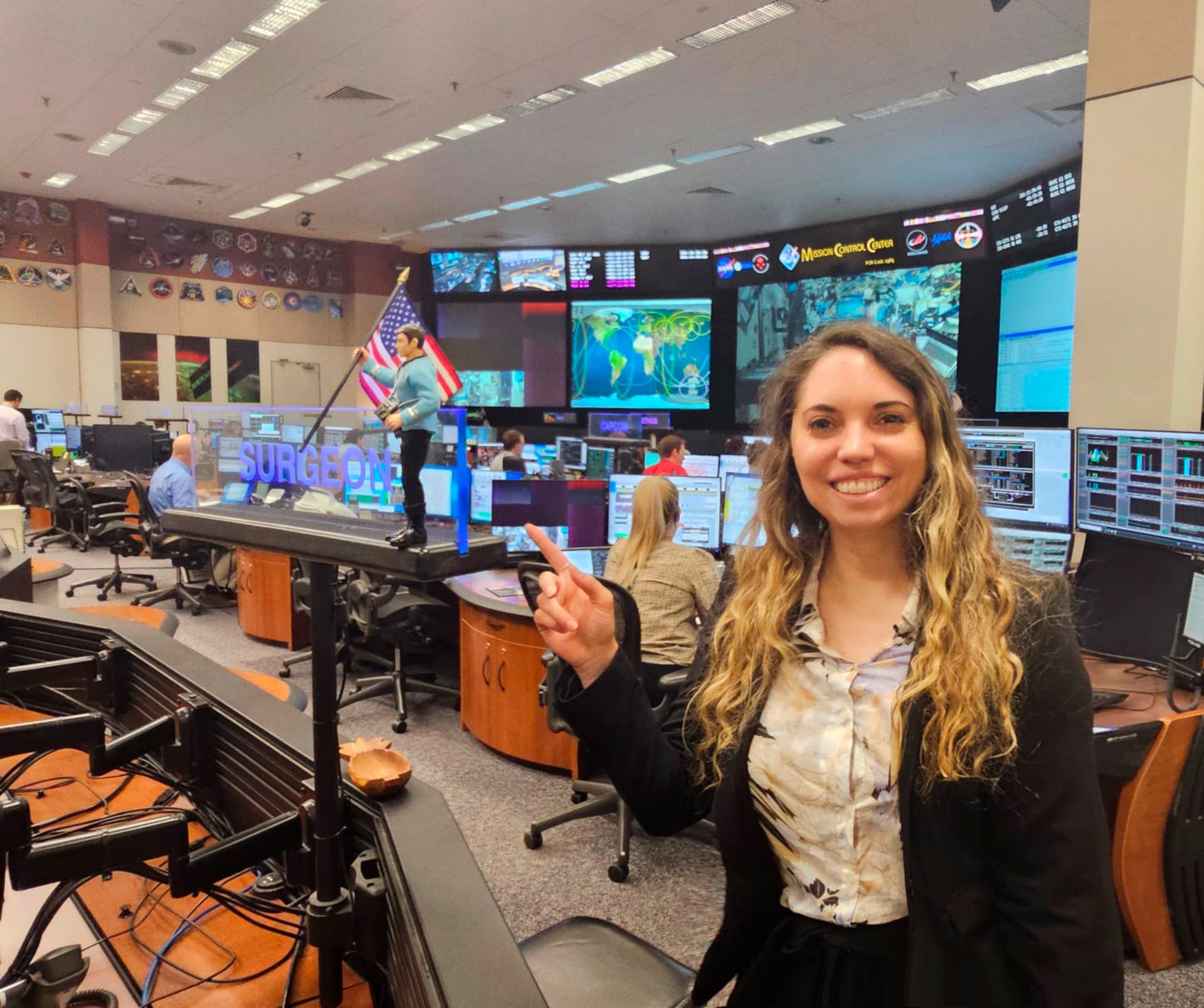Lorna Evans de la NASA sonríe y hace un signo de paz en el Centro de Control de Misiones, donde pantallas gigantes y una figura del Capitán Kirk son visibles.