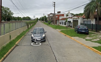 Calle asfaltada con dos coches estacionados, casas residenciales con jardín, un cerco perimetral, vías ferroviarias, postes de luz y cielo nublado