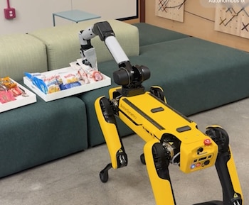 Un robot Spot de Boston Dynamics amarillo y negro con un brazo robótico gris, dos sofás verdes, dos bandejas blancas con paquetes de aperitivos variados, suelo gris