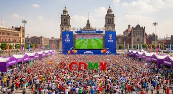 Gran multitud en el Zócalo de CDMX viendo un partido de fútbol en una pantalla gigante, con edificios históricos de fondo, confeti y carpas moradas.