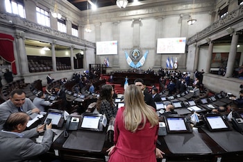 Un salón legislativo con curules de madera, alfombra roja, una bandera guatemalteca en primer plano y personas de traje
