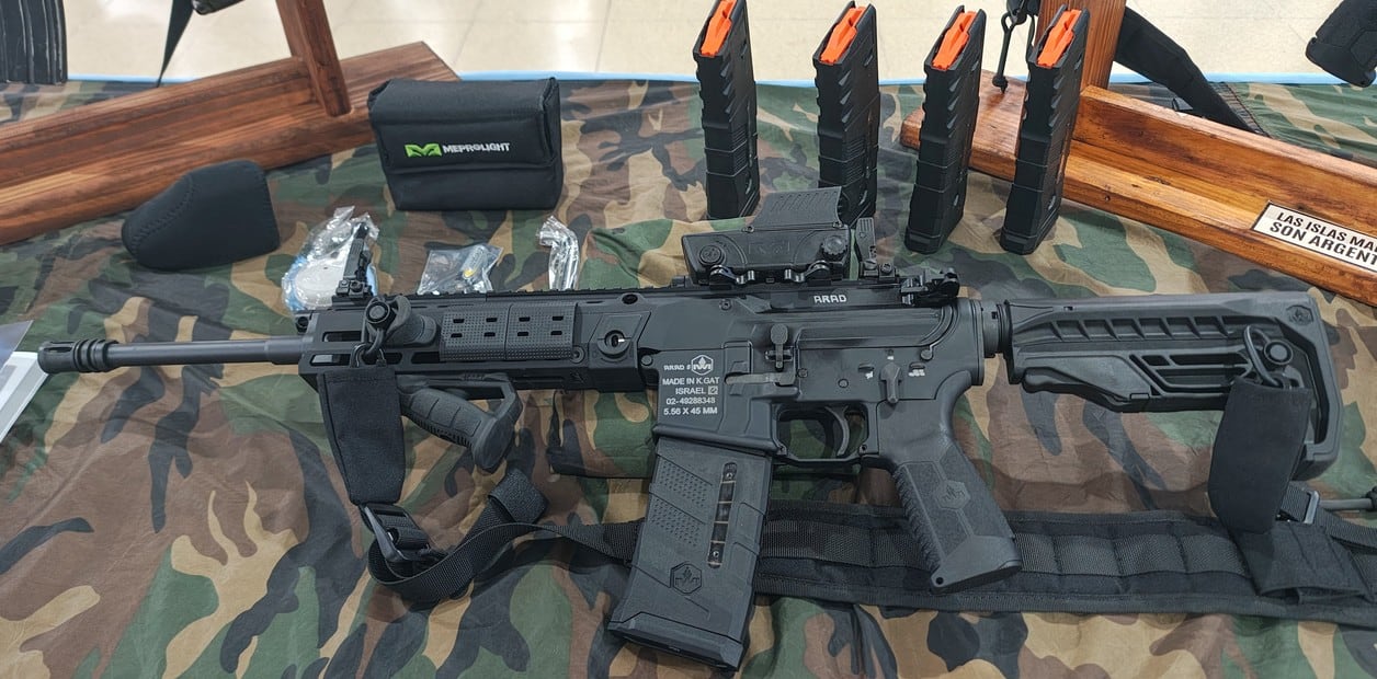 Un fusil de asalto IWI Arad de calibre 5,56 mm reposa sobre una tela de camuflaje junto a varios cargadores y accesorios militares en una exposición. (Ministerio de Defensa)