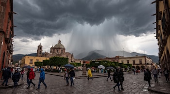 Nubes oscuras en el cielo, lluvia a la distancia, montañas, iglesia con cúpula, edificios, personas con paraguas y abrigos en calle empedrada.