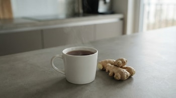Una taza de té blanco humeante y varias raíces de jengibre están sobre una mesada de cocina gris, con el fondo de una cocina moderna difuminado.