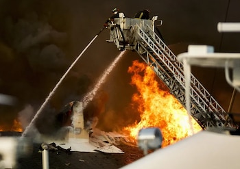 Vista aérea de bomberos en una escalera elevadora extinguiendo un gran incendio con llamas y humo espeso en un edificio. Dos chorros de agua impactan las llamas