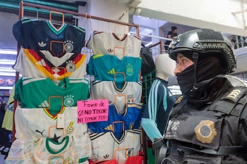 Un policía de la SSC con casco y uniforme táctico mira a la derecha, de pie junto a un exhibidor de camisetas deportivas, algunas con logotipos de equipos de fútbol y una señal de "no tocar"