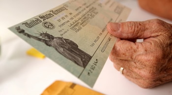 Mano de una persona mayor que sostiene un cheque impreso del Tesoro de Estados Unidos junto a un sobre en una mesa.